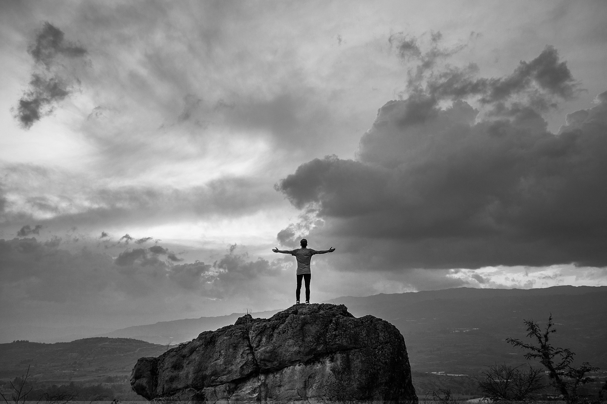 A man standing on top of a large rock with his arms spread wide under a stormy sky