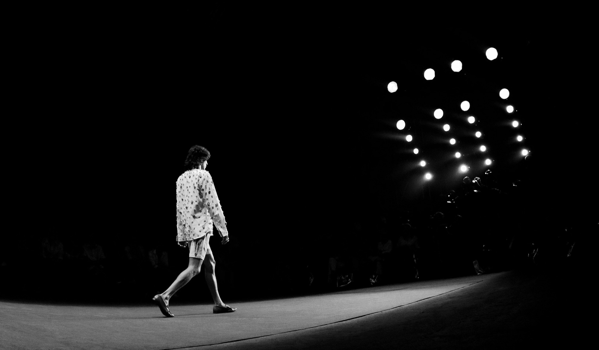 A model walks a dark catwalk towards a curve of bright stage lights